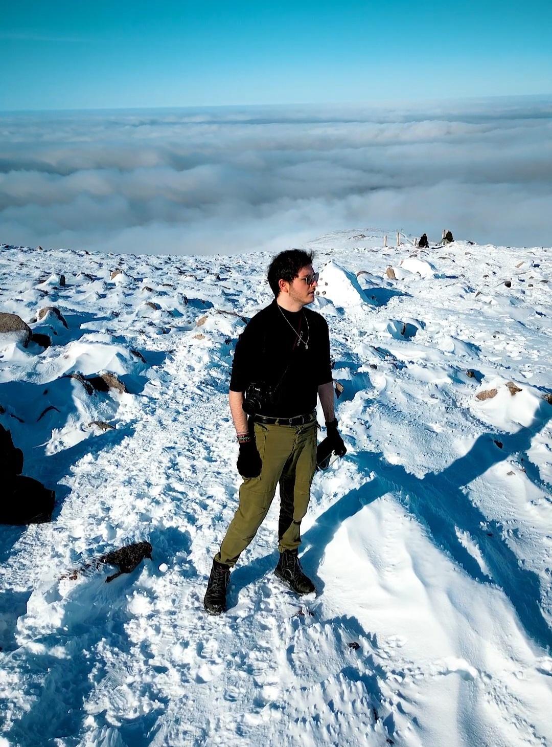 Ruben Bitoun, appareil photo en bandoulière, au sommet d'une montagne enneigée au-dessus d'une mer de nuages